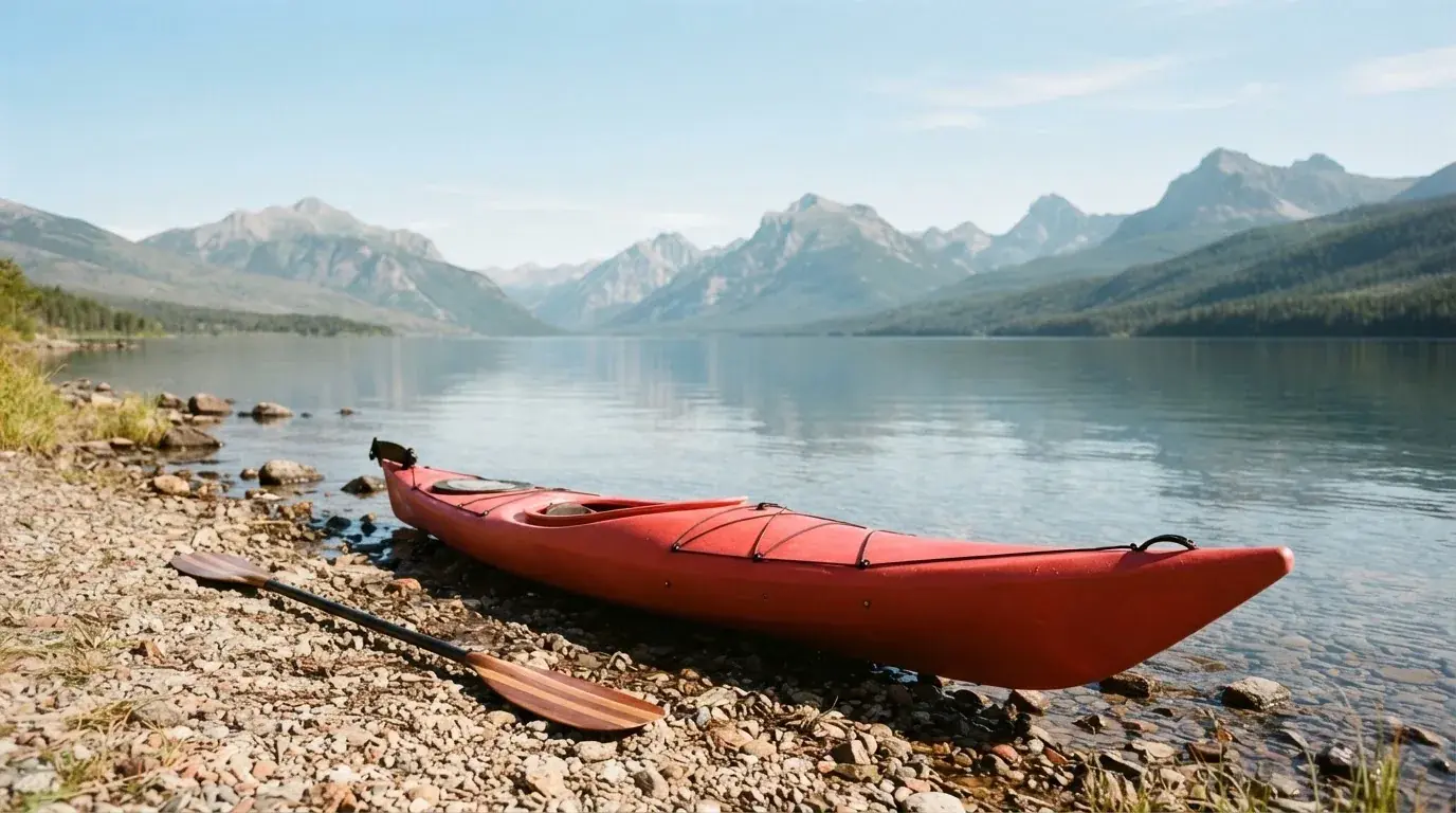 Ein Kajak liegt am Ufer eines Bergsees, daneben ein Paddel, das Wasser ist glatt und der Himmel klar.