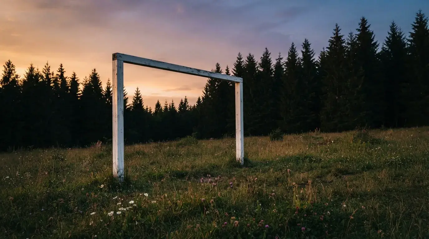 Ein Fussballtor ohne Netz steht auf einer Wiese am Waldrand, die Abenddämmerung färbt den Himmel orange.