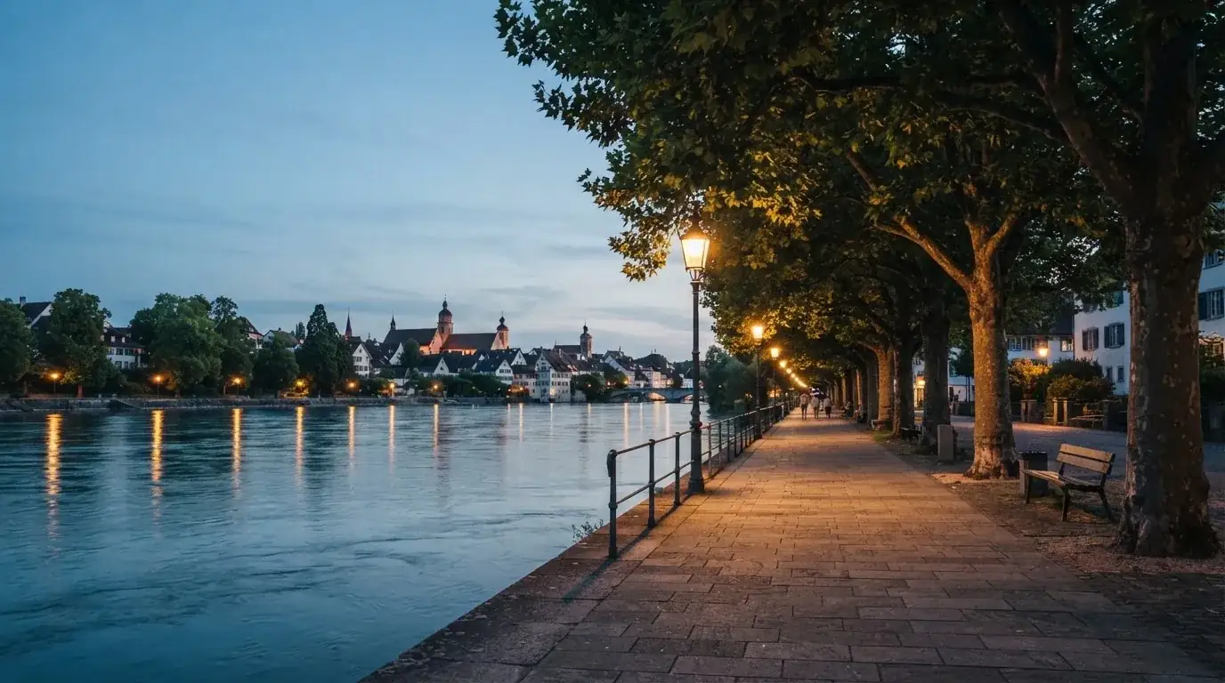 Eine Rheinpromenade mit hohen Bäumen am Abend, das Wasser spiegelt das Licht der ersten Laternen.