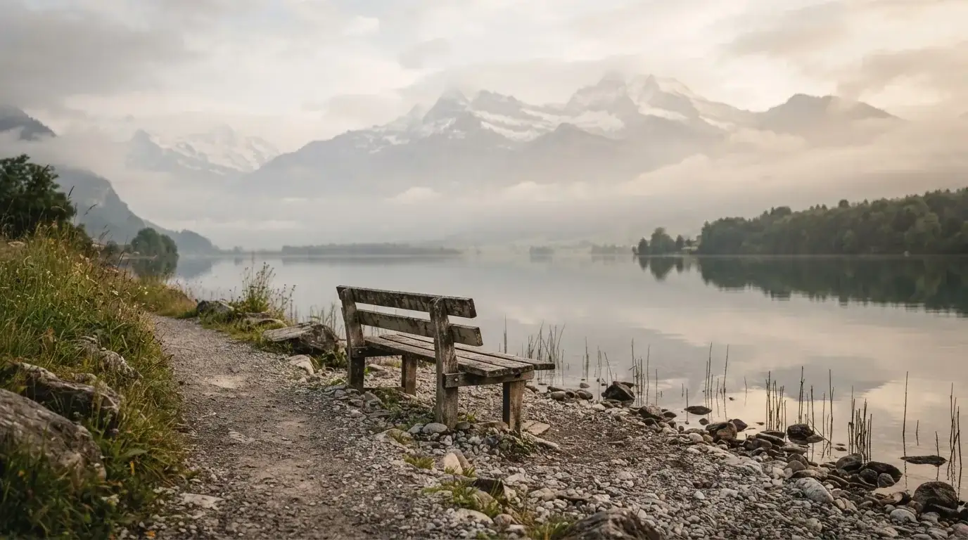 Eine Holzbank an einem Seeufer mit Blick auf eine Bergkette im Morgendunst, das Wasser ist spiegelglatt.