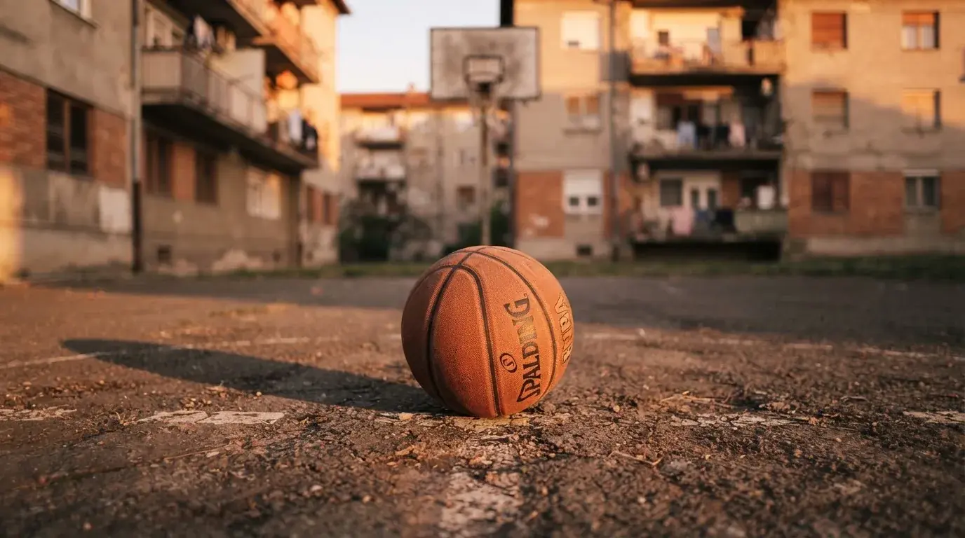 Ein Basketball liegt auf einem Asphaltplatz in einem Hinterhof zwischen Wohnblöcken, das Abendlicht färbt die Wände warm.