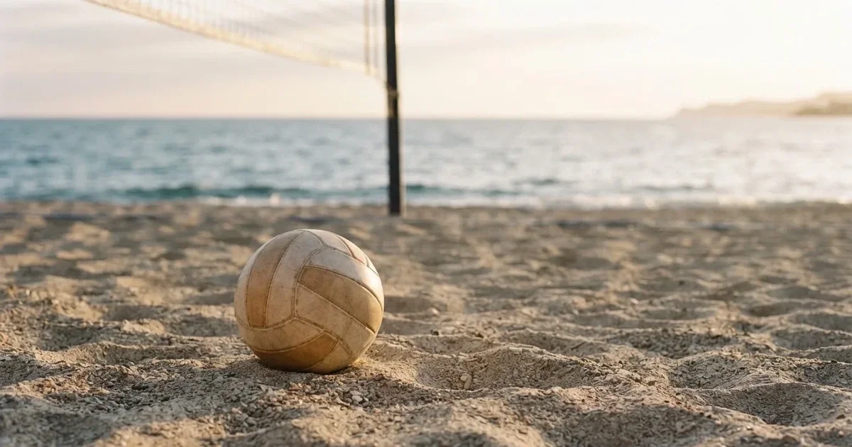 Ein Volleyball liegt auf feinem hellem Sand, ein gespanntes Netz verschwimmt dahinter, das Licht kommt von einem weiten Meer.