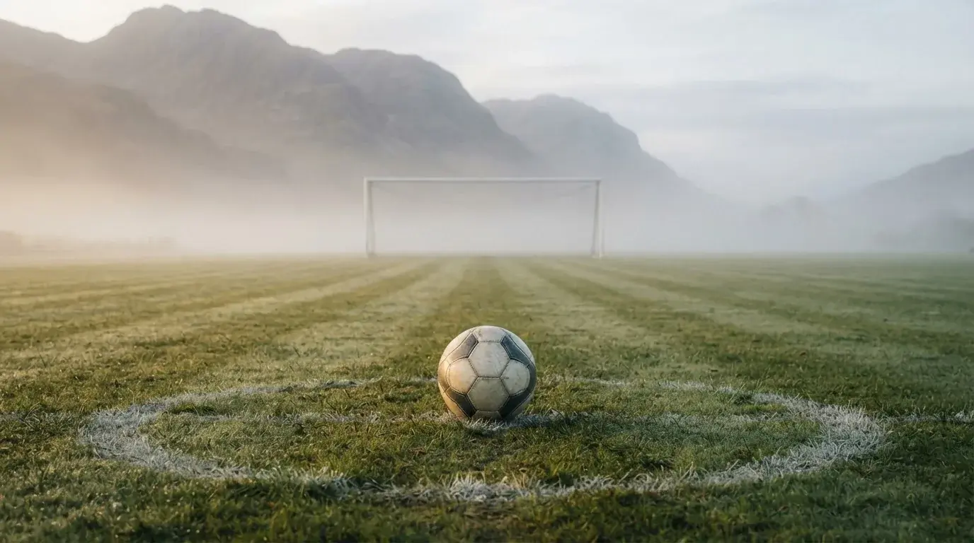 Ein einzelner Fussball auf dem Mittelpunkt eines leeren Rasens im Morgennebel, hinter dem Tor ragen Berge auf