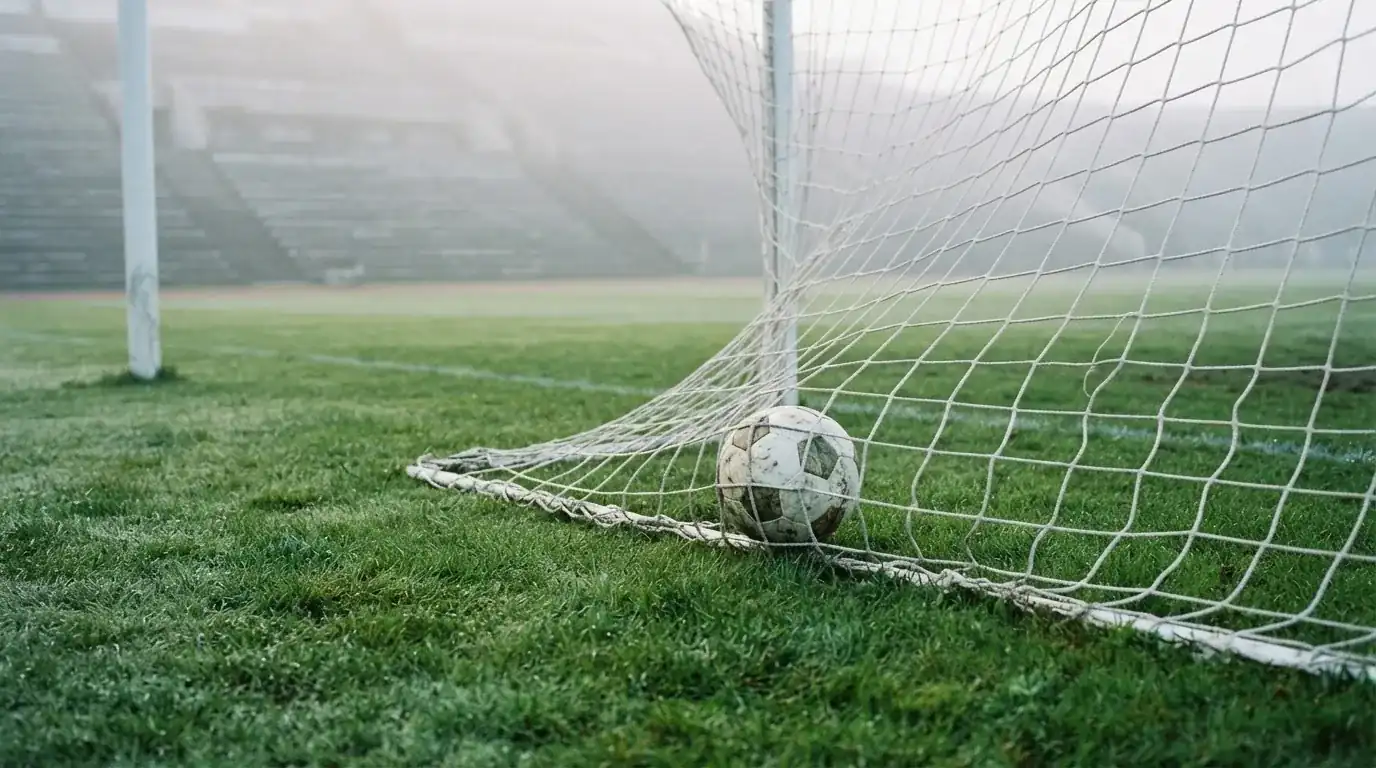 Ein Fussball liegt im Netz hinter einem Tor, weicher Morgennebel hängt über dem Rasen