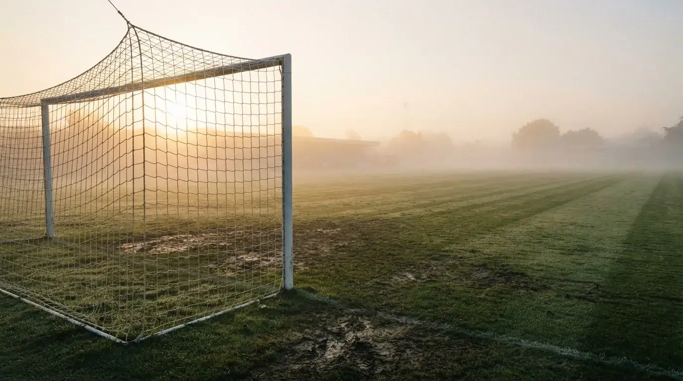 Ein Torpfosten mit Netz steht im Gegenlicht auf einem leeren Fussballplatz, über dem Rasen liegt Nebel.